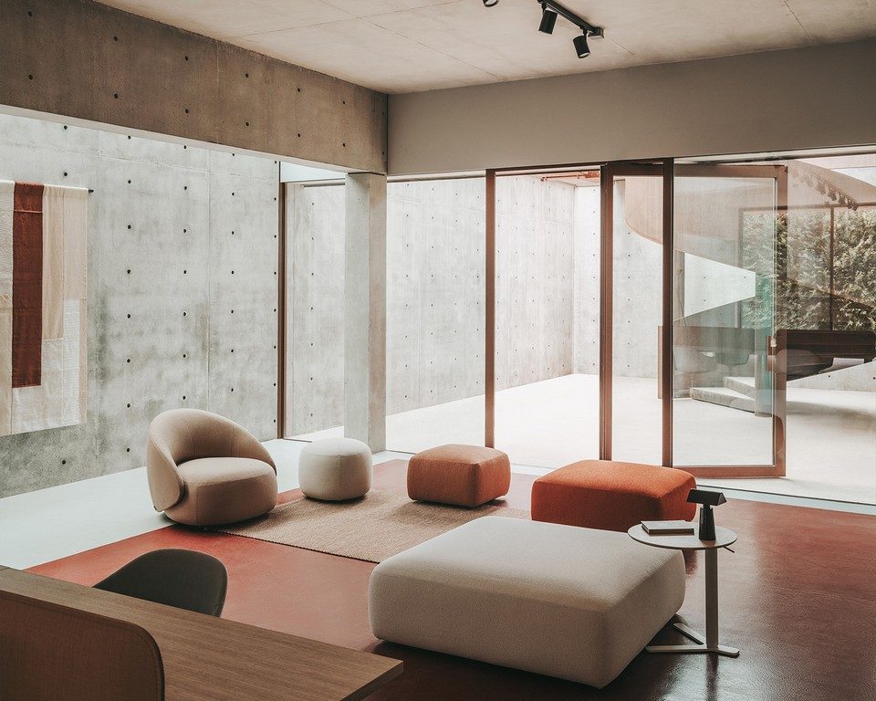 Zone d'attente contemporaine avec sol en béton ciré rouge brique présentant un fauteuil club KIPU, un grand pouf carré blanc cassé au premier plan et trois poufs galets déclinés dans des teintes terracotta et ocre devant une grande baie vitrée.
