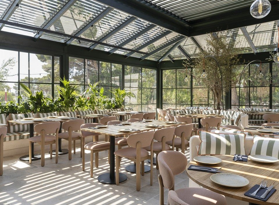 Salle de restaurant lumineuse aménagée sous une structure en verrière avec des chaises en bois clair et assises roses BAOBAB.