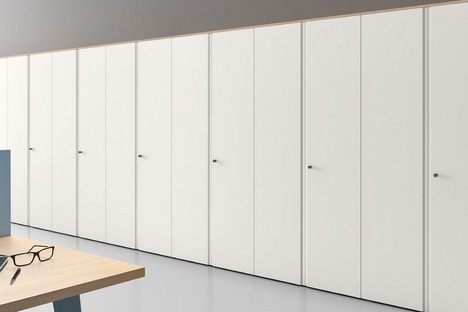 Perspective sur un grand placard de rangement de bureau GALAXY composé d'un alignement de portes battantes blanches et minimalistes avec de discrets boutons de poignées ronds. Au premier plan, un angle de table de réunion ou de bureau en bois clair accueille une paire de lunettes, illustrant la parfaite intégration visuelle de ce système d'armoire modulaire dans un espace de travail épuré.