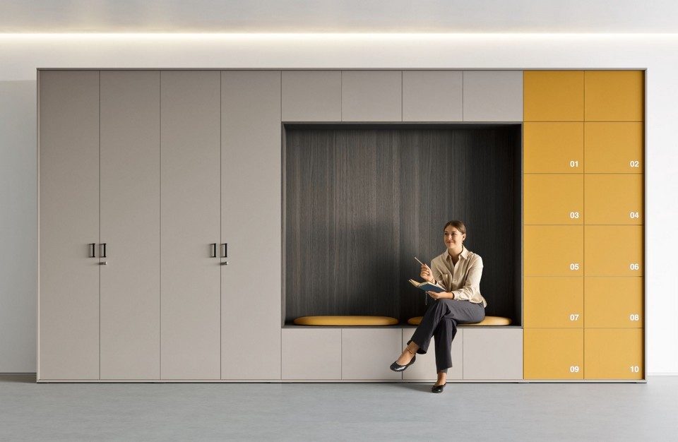 Vue de face de l'armoire casier personnalisable GALAXY structurant un espace de travail. Ce système de rangement modulaire combine des placards de bureau gris clair à gauche, une alcôve centrale en bois foncé avec banquette intégrée occupée par une collaboratrice, et un bloc de dix casiers individuels de couleur jaune moutarde numérotés pour le flex-office.