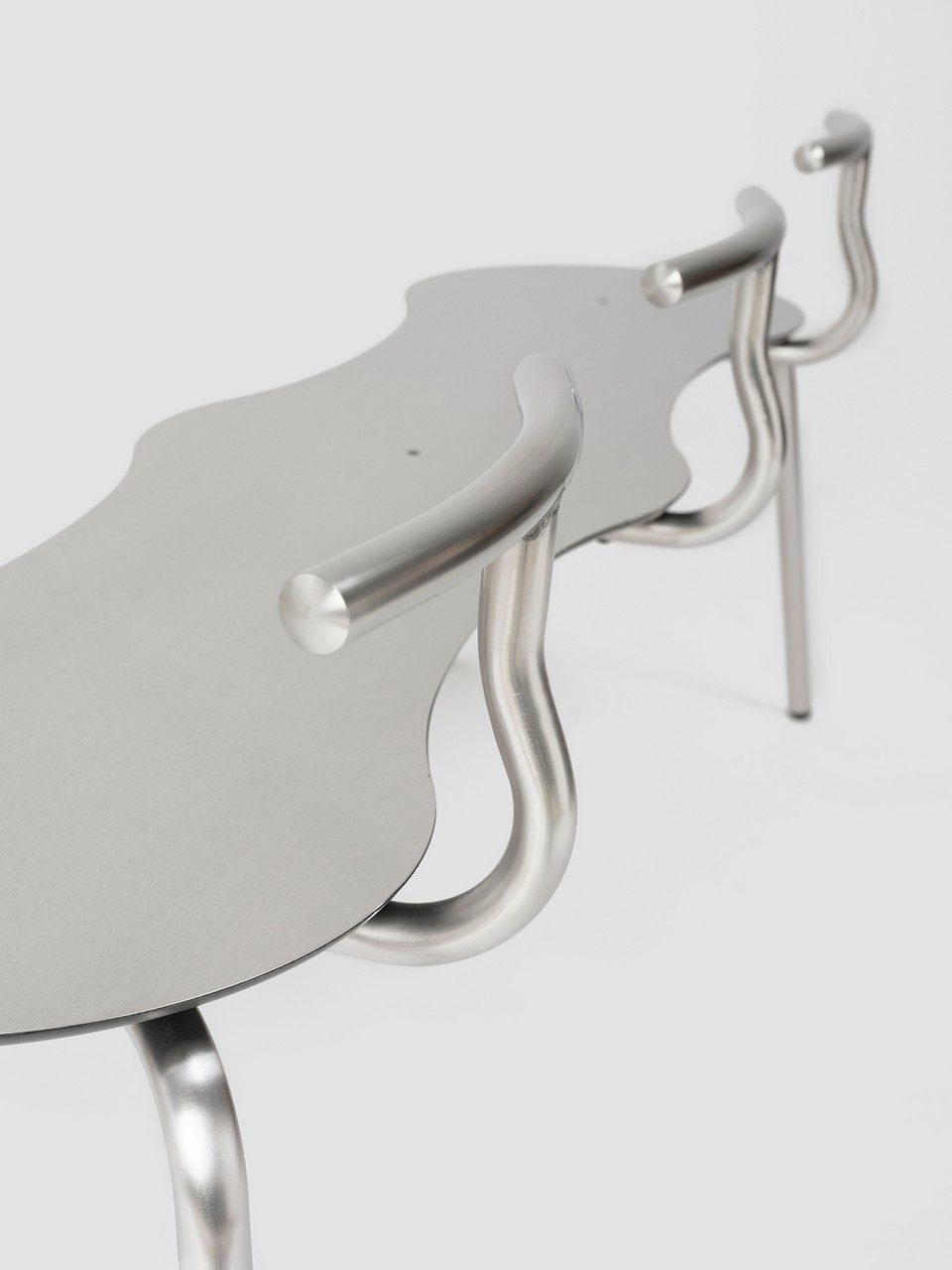 Gros plan en perspective du banc triple en acier inoxydable TAM par Ondarreta, montrant l'assise ondulée en inox brossé et ses trois dossiers tubulaires courbés amovibles. Mobilier d'assise minimaliste haut de gamme et tendance chrome pour terrasse de restaurant ou espace lounge.