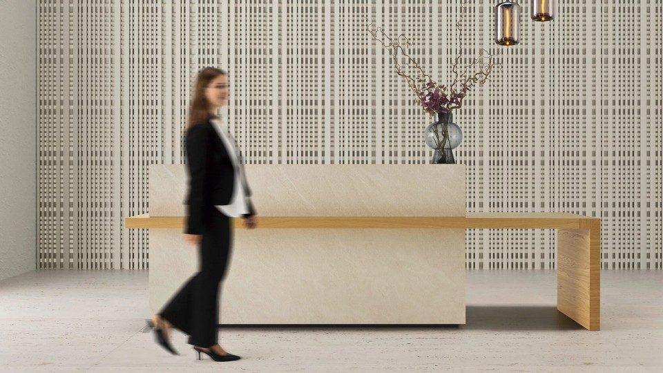 Banque d'accueil monolithique design Kyo avec structure minérale beige BioArgilla et plateau en placage bois étendu en aile libre. L'image montre une femme floue marchant devant le comptoir dans un hall d'accueil contemporain doté d'un mur acoustique texturé et de luminaires design. L'éclairage LED continu au socle souligne la texture minérale et l'architecture épurée, créant une atmosphère haut de gamme et feutrée pour un coworking ou un hôtel-boutique.