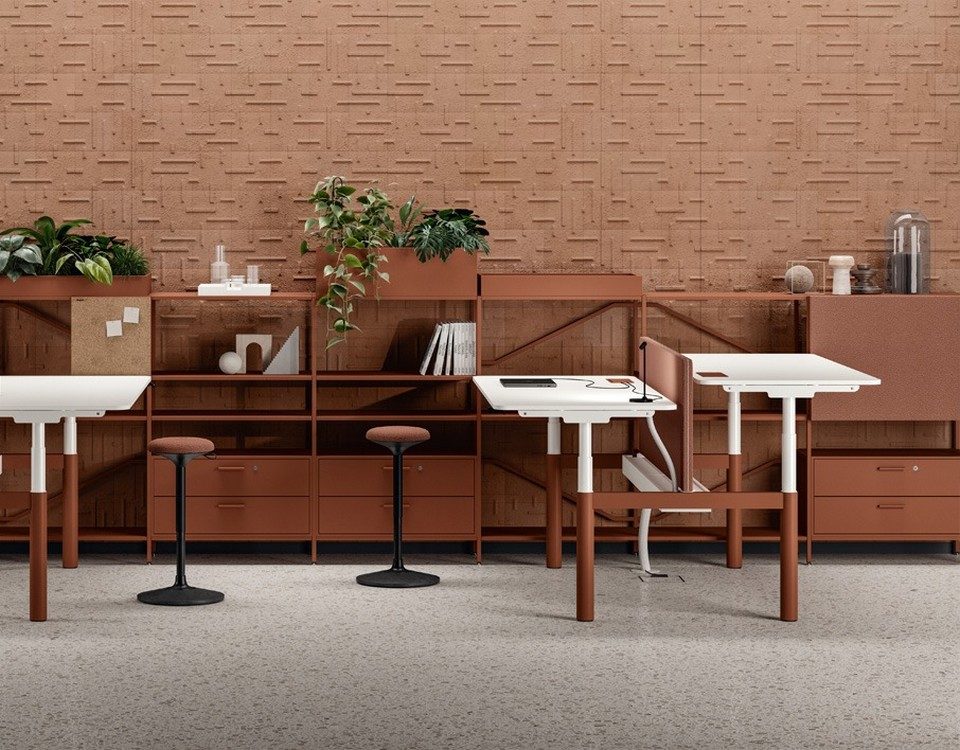Bureaux assis-debout individuels réglables en hauteur NUVOLA de Bralco avec piétement métallique terracotta et plateau blanc, disposés en rangée devant une grande étagère de rangement assortie dans un open space.