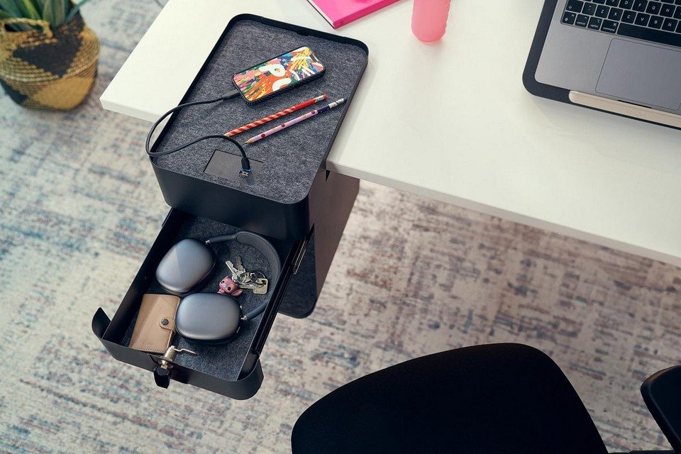 Vue de dessus en situation de travail d'un caisson suspendu BENTO noir avec son tiroir ouvert révélant des accessoires personnels, un smartphone en charge sur le plateau supérieur en feutre gris grâce aux prises USB intégrées, fixé à un bureau blanc.