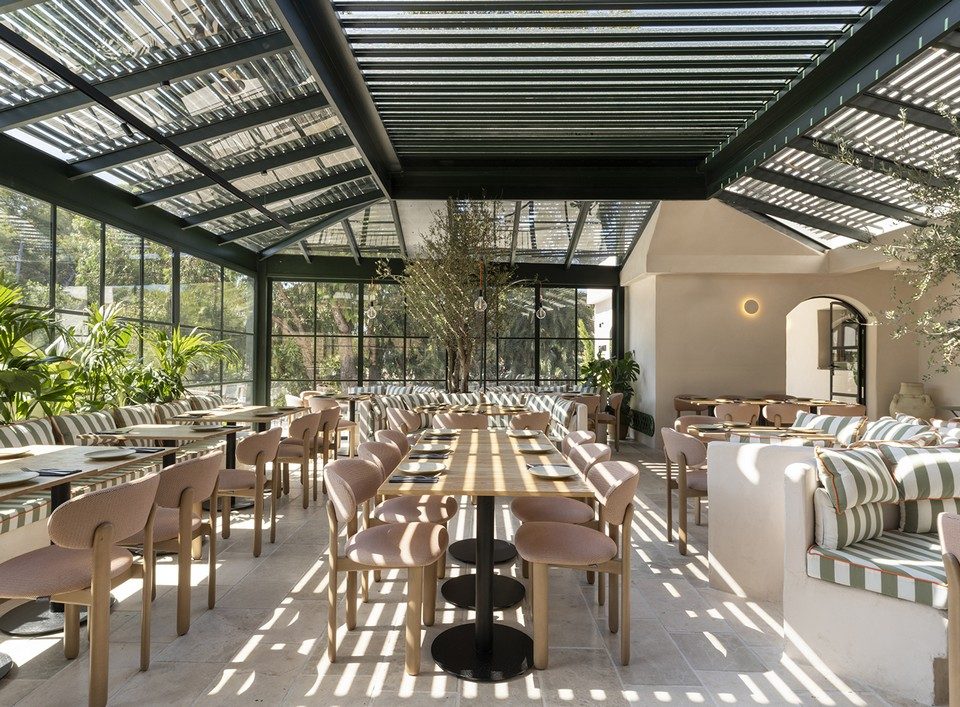 Aménagement d'une salle de restaurant sous verrière avec chaises en bois clair et revêtement rose BAOBAB.