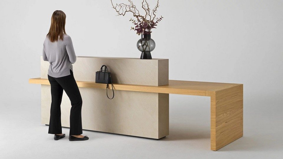 Vue de trois-quarts d'une femme debout de dos devant la banque d'accueil Kyo en finition minérale beige BioArgile. Un sac à main noir est posé sur l'épais plateau en bois qui se prolonge en aile libre sur la droite. Un vase en verre fumé décore la rehausse supérieure.