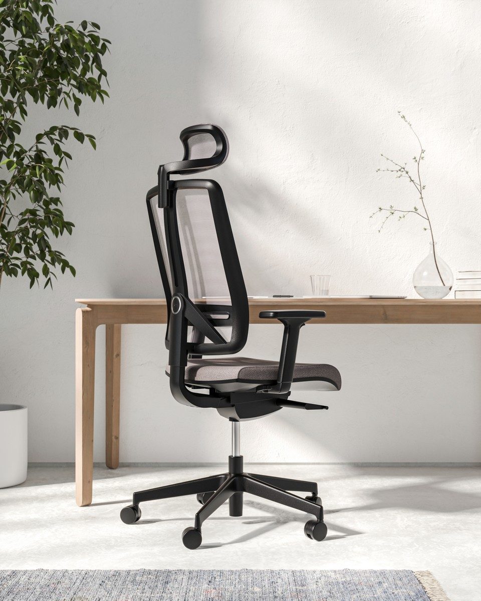 Vue de trois-quarts arrière du fauteuil de bureau ergonomique FLEXI avec structure noire, dossier en maille respirante, têtière réglable et assise grise, placé devant un bureau minimaliste en bois clair.