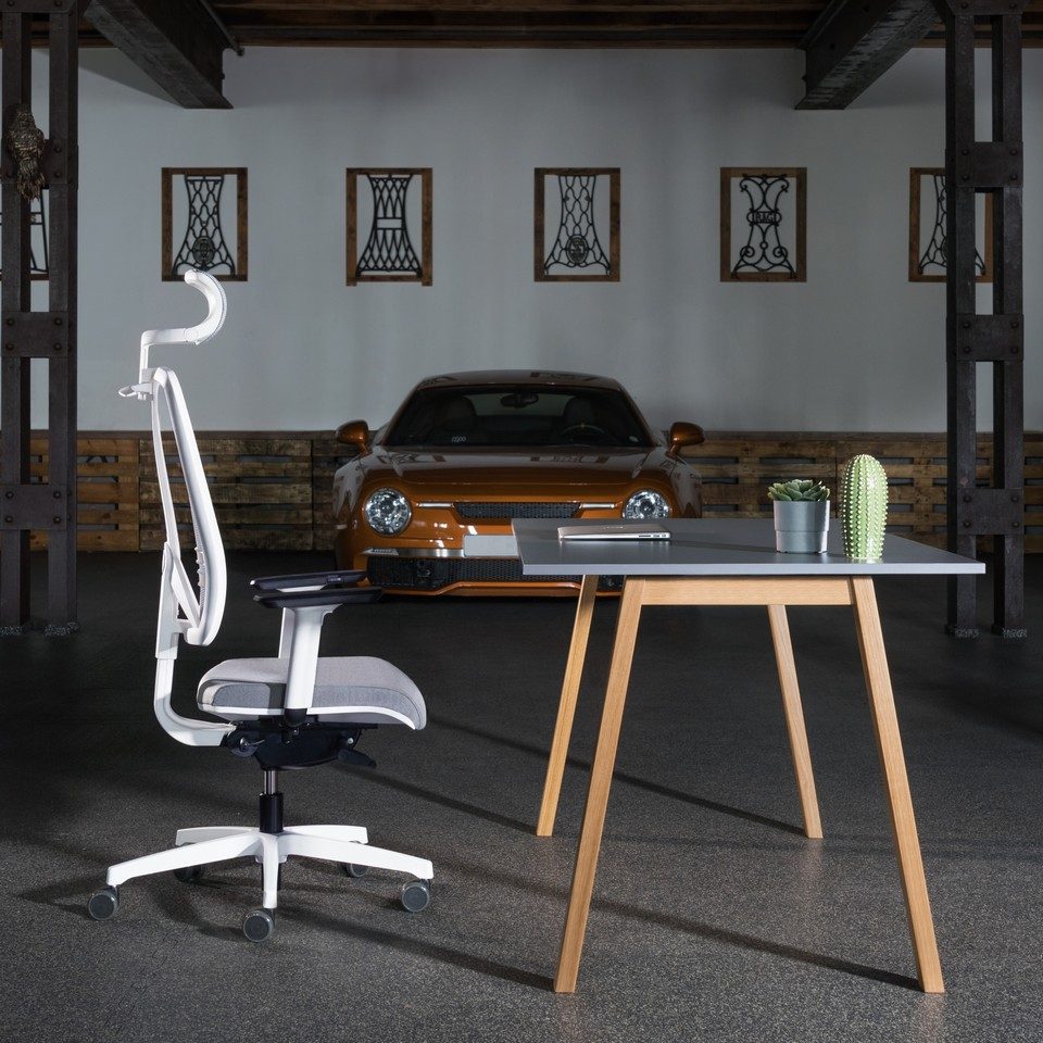 Vue de profil du fauteuil de bureau ergonomique FLEXI blanc et gris avec têtière, placé devant un bureau moderne en bois dans un espace au style industriel avec une voiture de sport en arrière-plan.