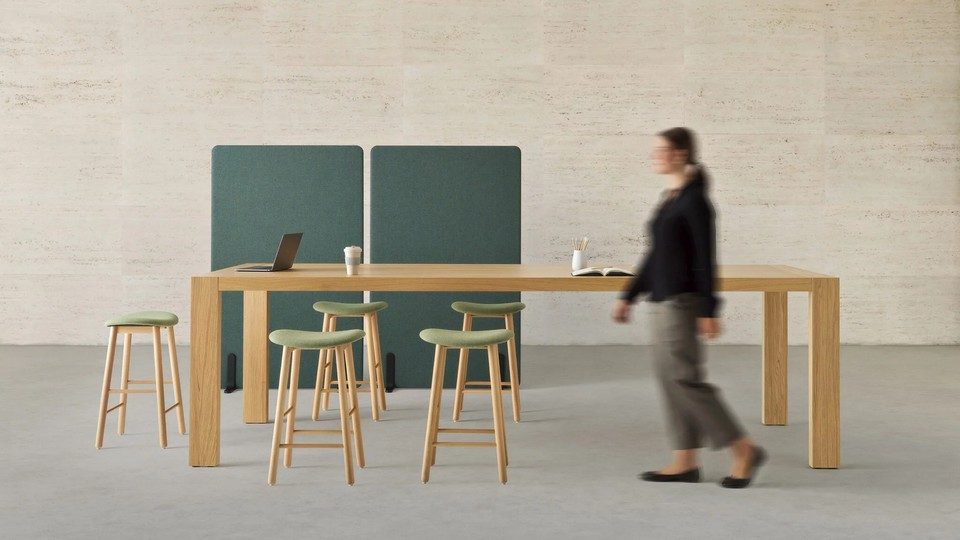 Grande table haute de coworking Kyo en placage bois avec plusieurs tabourets de bar aux assises vertes. En arrière-plan, deux cloisons acoustiques mobiles vertes délimitent l'espace de réunion au sein d'un bureau collaboratif moderne et épuré. Une personne passe au premier plan.