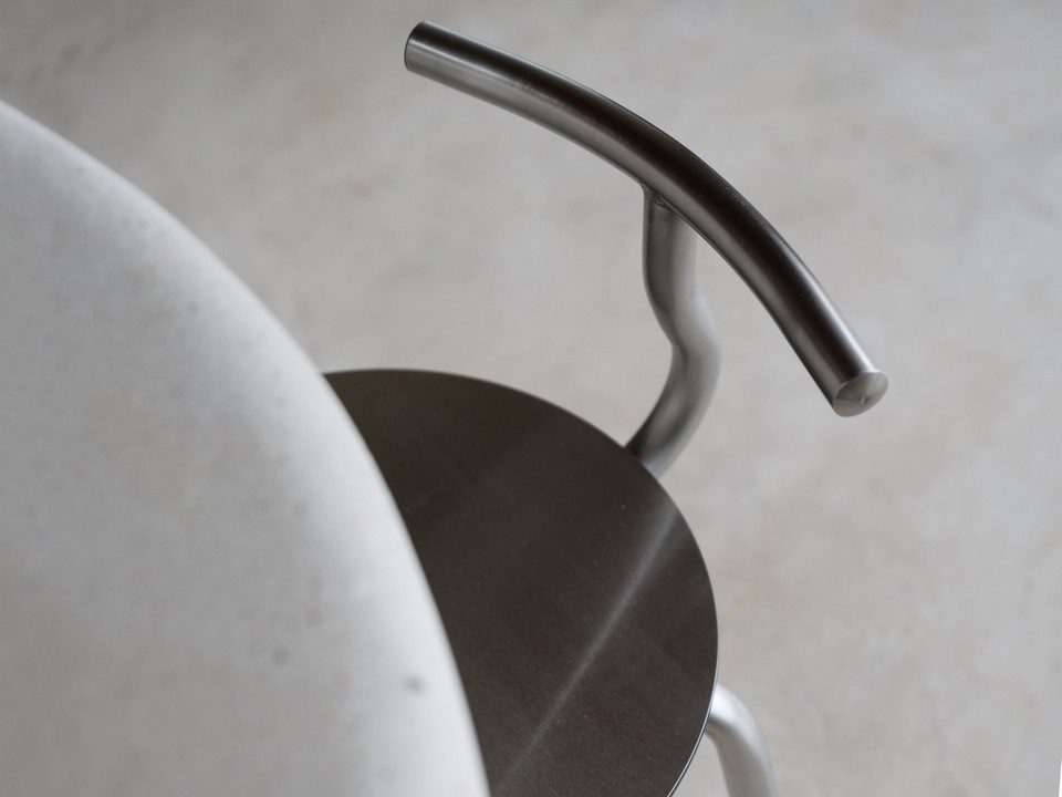 Gros plan aérien sur le dossier tubulaire en acier inoxydable brossé et l'assise circulaire du tabouret TAM par Ondarreta, glissé sous une table. Détail du design minimaliste et de la finition métallique tendance chrome pour l'agencement de restaurant style bistrot pro.