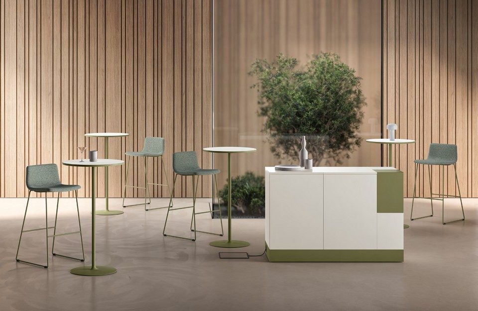 Station café autonome MAG blanche et vert olive installée dans une cafétéria d'entreprise moderne. Le meuble tisanerie est entouré de tables mange-debout et de tabourets de bar assortis, devant un mur en tasseaux de bois. Mobilier professionnel pour l'aménagement d'espaces de pause et de convivialité au bureau.