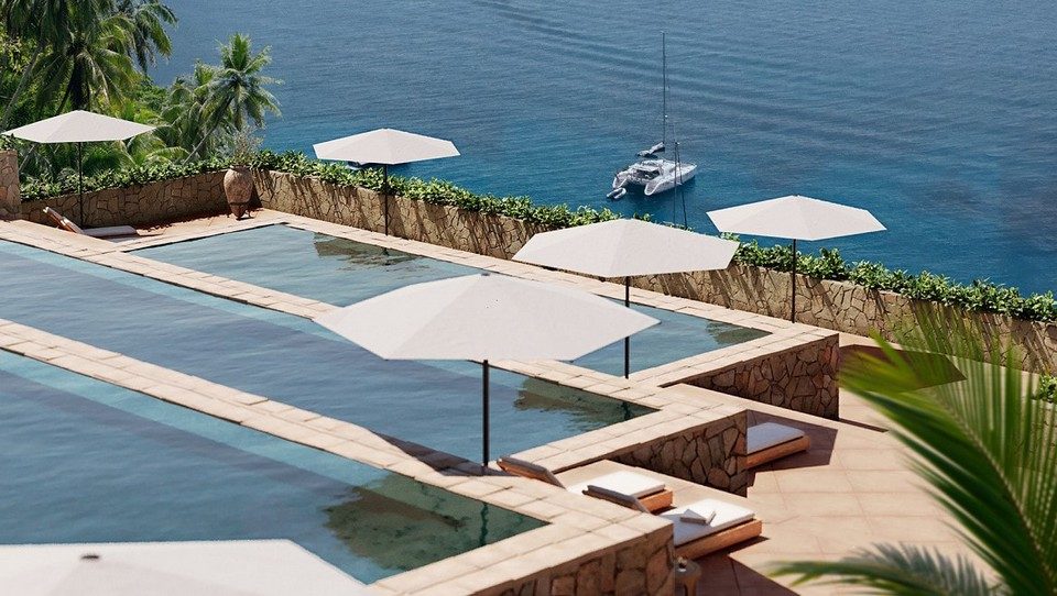 Alignement de parasols NATA en bord de piscine à débordement avec vue panoramique sur la mer et un yacht au mouillage.