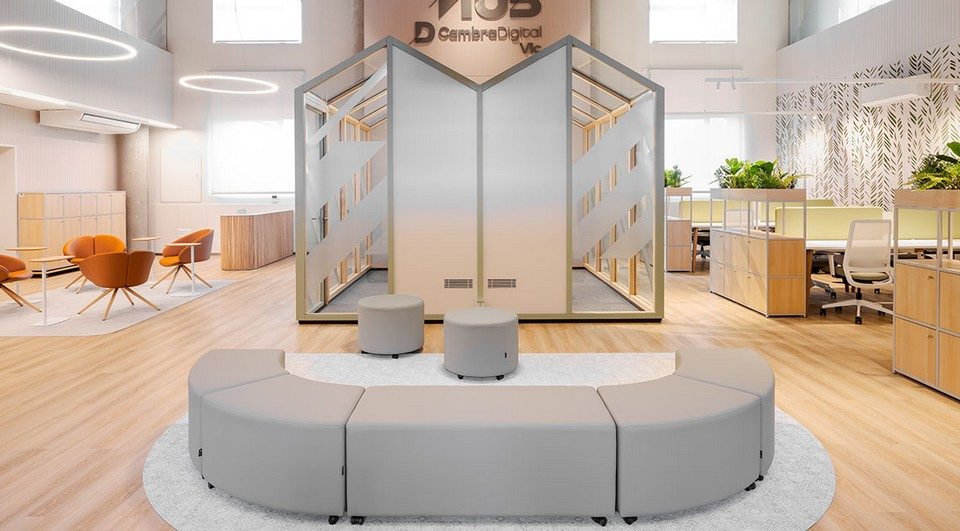 Pavillon de réunion de coworking EVA en configuration double toit en pointe avec vitrages sérigraphiés, positionné au centre d'un grand espace de travail collaboratif avec un canapé modulaire courbe au premier plan. Aménagement industriel chic.