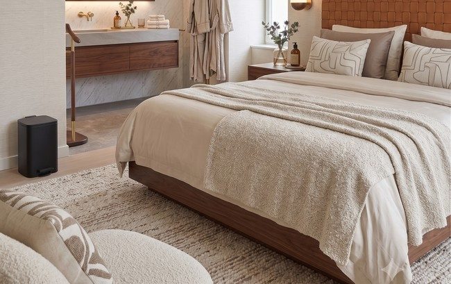 Vue d'ensemble d'une chambre d'hôtel haut de gamme montrant une poubelle à pédale noire rectangulaire posée sur un tapis texturé au pied d'un lit en bois, avec une salle de bain ouverte en arrière-plan. Équipement de propreté discret et esthétique pour l'hôtellerie.