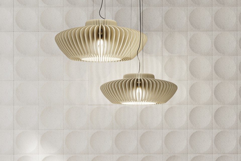 Deux suspensions d'éclairage acoustique design RAYA en lamelles de feutre de PET recyclé beige. Les luminaires diffusent une lumière chaude devant un revêtement mural composé de dalles acoustiques blanches à reliefs circulaires. Solution d'insonorisation et de confort visuel pour l'aménagement de bureaux tertiaires ou d'espaces lounge.