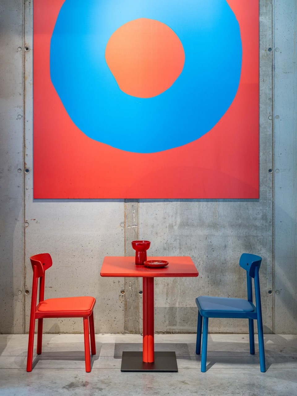 Table ONE rouge et chaises design bleues devant un mur en béton brut pour un style urbain.