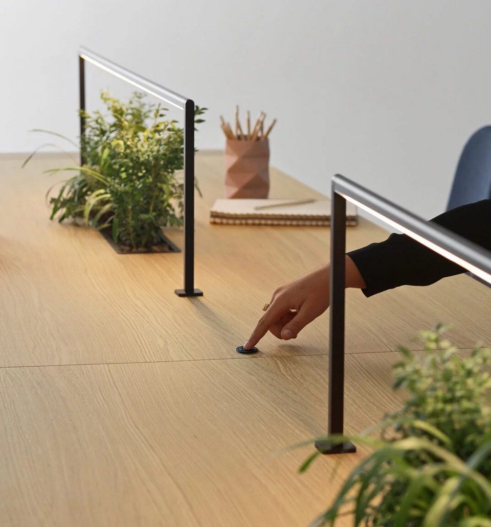Gros plan sur le plateau en placage bois d'une table de coworking Kyo. Une main actionne un bouton-poussoir affleurant et intégré dans le bois pour allumer des lampes LED longilignes. Des jardinières centrales avec des plantes vertes sont visibles en arrière-plan.