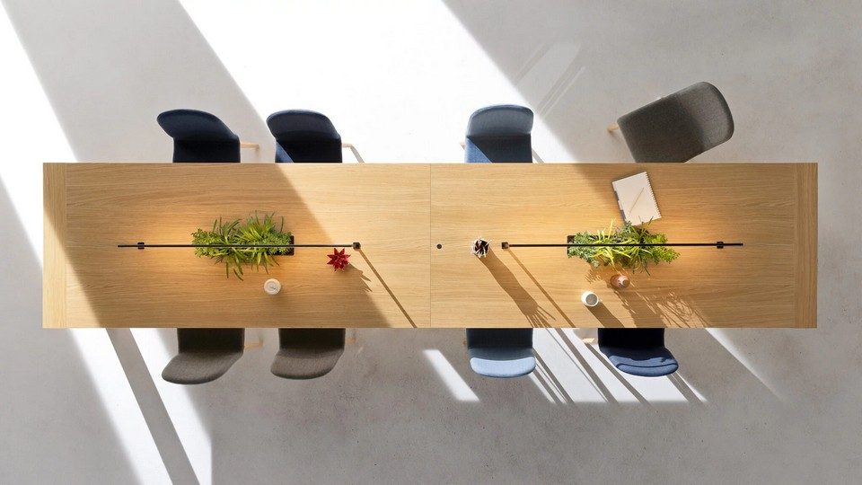Vue de dessus d'une grande table de coworking monolithique Kyo en placage bois clair. Elle dispose de jardinières centrales intégrées avec des plantes vertes et des luminaires LED fins et longilignes. Huit chaises de bureau modernes dans des tons de bleu et de gris sont disposées autour. Le sol en béton poli présente des motifs d'ombres et de lumière du soleil.