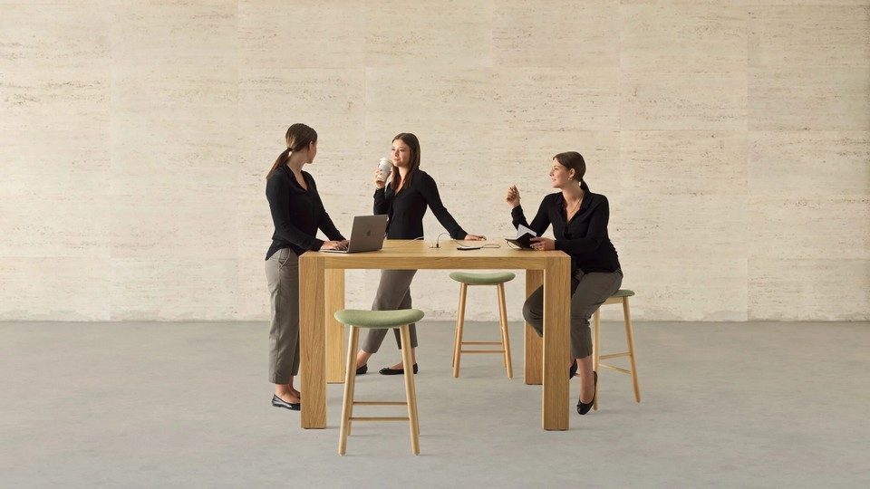 ne photo d'ambiance montrant trois femmes collaborant et socialisent autour de la grande table de coworking haute Kyo (finition bois véritable Rovere Classico) de 950 mm. Elles utilisent un ordinateur portable, un carnet et une tasse. Autour d'elles, des tabourets de bar aux assises vert clair. L'arrière-plan présente un grand mur texturé en travertin et un sol en béton. Le mobilier est monolithique et épuré, évoquant une esthétique high-tech.