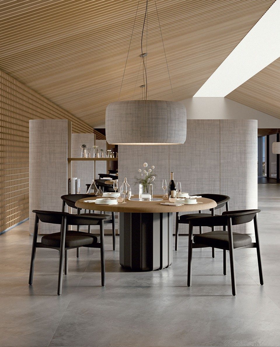 Table d'hôte ronde de design SABREE avec plateau en bois de frêne teinté chêne et piètement colonne central flûté anthracite, entourée de chaises noires dans un restaurant gastronomique ou un espace collaboratif haut de gamme.