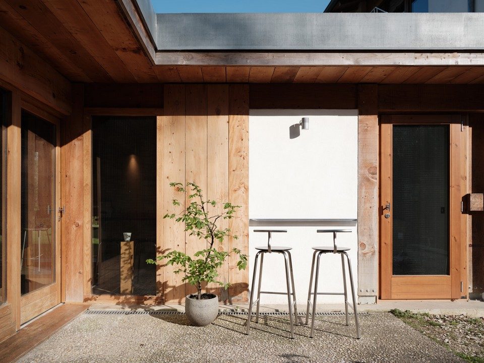 Deux tabourets de bar hauts en inox brossé avec dossier amovible TAM par Ondarreta installés sur la terrasse en bois d'un café contemporain. Mobilier outdoor minimaliste style bistrot en acier inoxydable pour l'aménagement extérieur d'hôtels et restaurants.